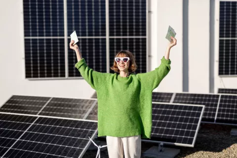 Eine Frau in grünem Pullover steht vor ihrer Photovoltaikanlage und hält ihre Arme nach oben, während sie in den Händen Geldscheine hält.